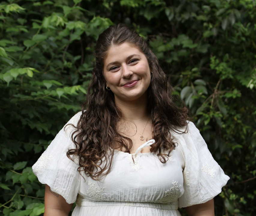 A young woman with long, curly brown hair smiling outdoors in front of green foliage, wearing a white blouse and gold jewelry.