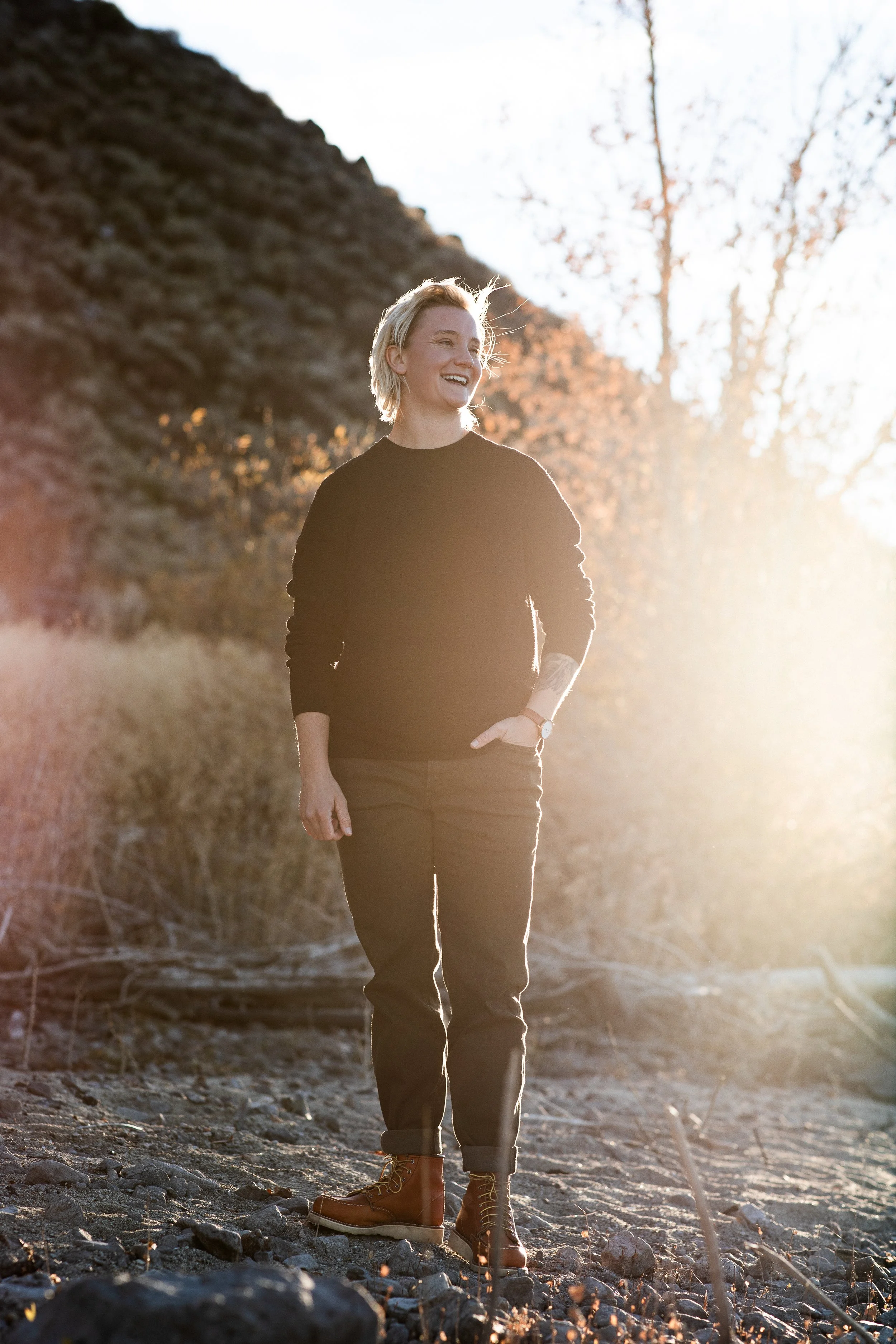 A woman with short blonde hair, wearing a black sweater, brown pants, and brown boots, standing outdoors on rocky terrain with a mountain and leafless trees in the background, smiling as sunlight shines behind her.