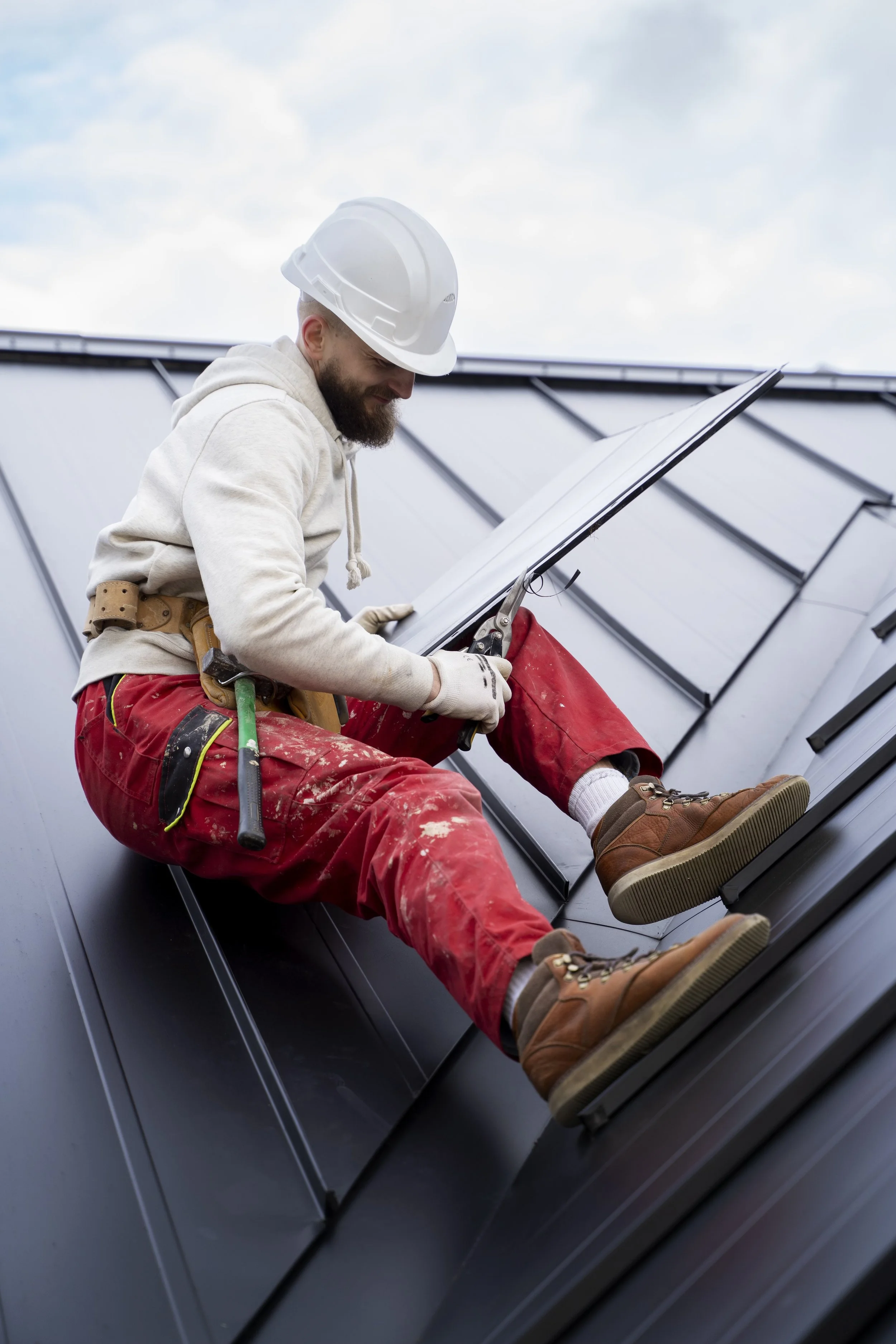 A construction worker wearing a white hard hat, beige hoodie, red work pants, and brown work boots installing or inspecting solar panels on a metal roof.
