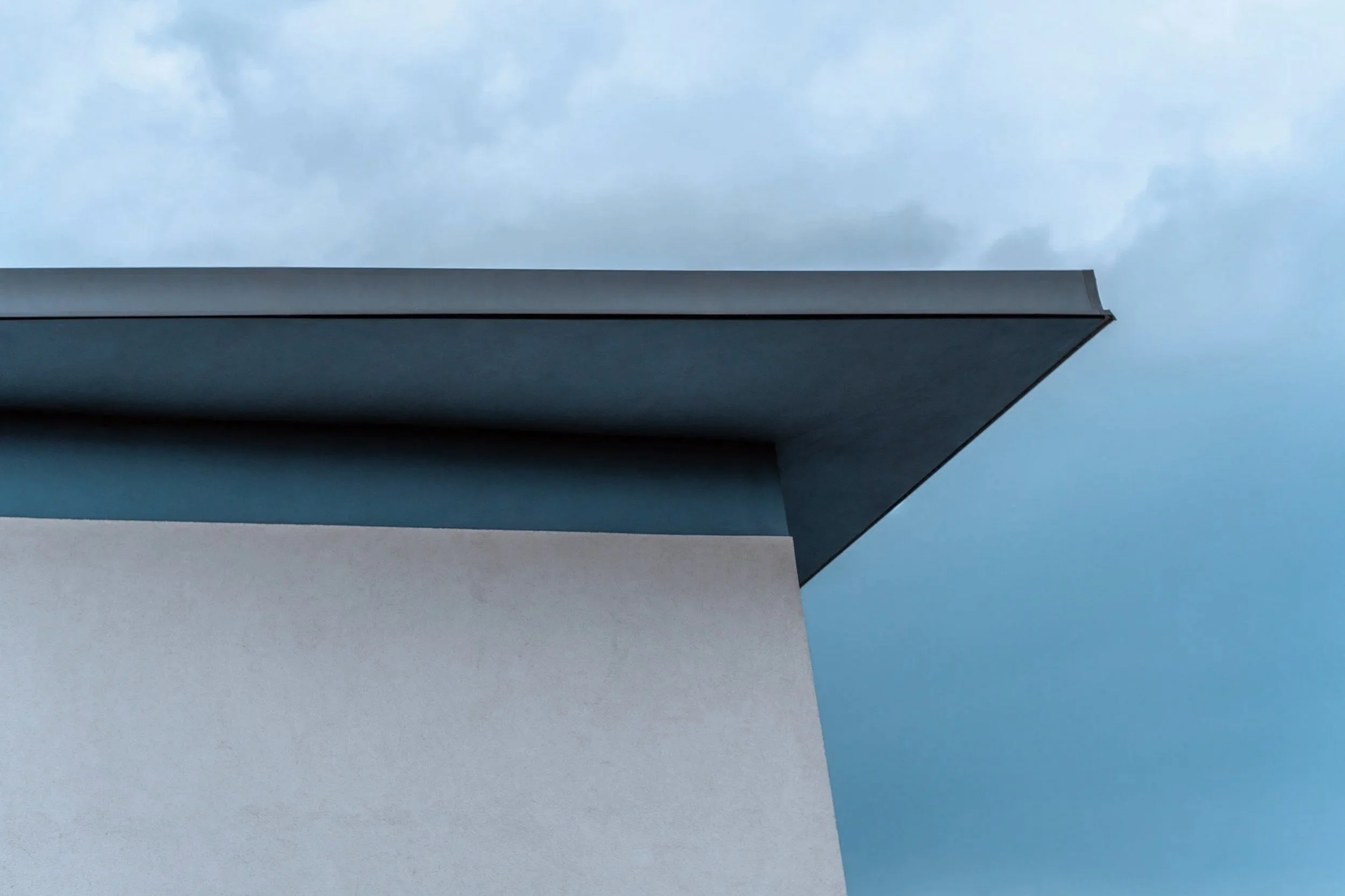 Close-up of a modern building's corner with a white wall and a dark overhang against a cloudy sky.
