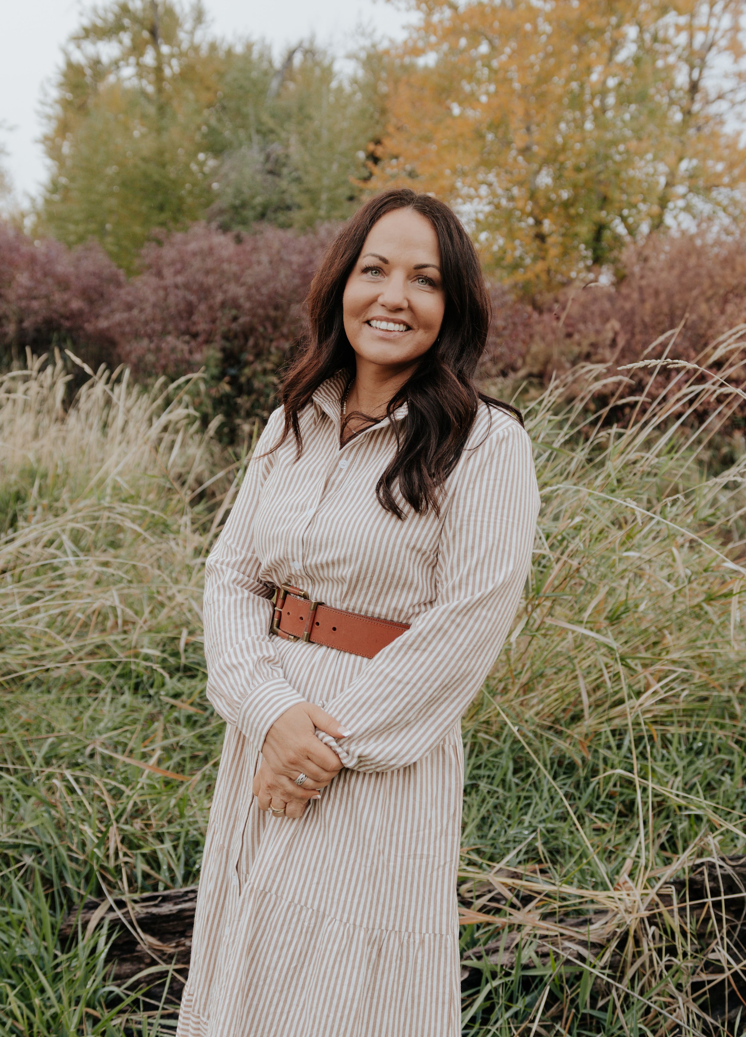 A woman with long dark hair, light-colored eyes, and a striped beige and white dress standing outdoors in a natural setting with fall foliage, trees with autumn leaves, and tall grasses.