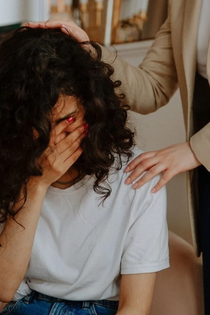A woman with curly dark hair covering her face with her hand, seated indoors. Another person with light skin, wearing a tan blazer, is placing a comforting hand on her shoulder.