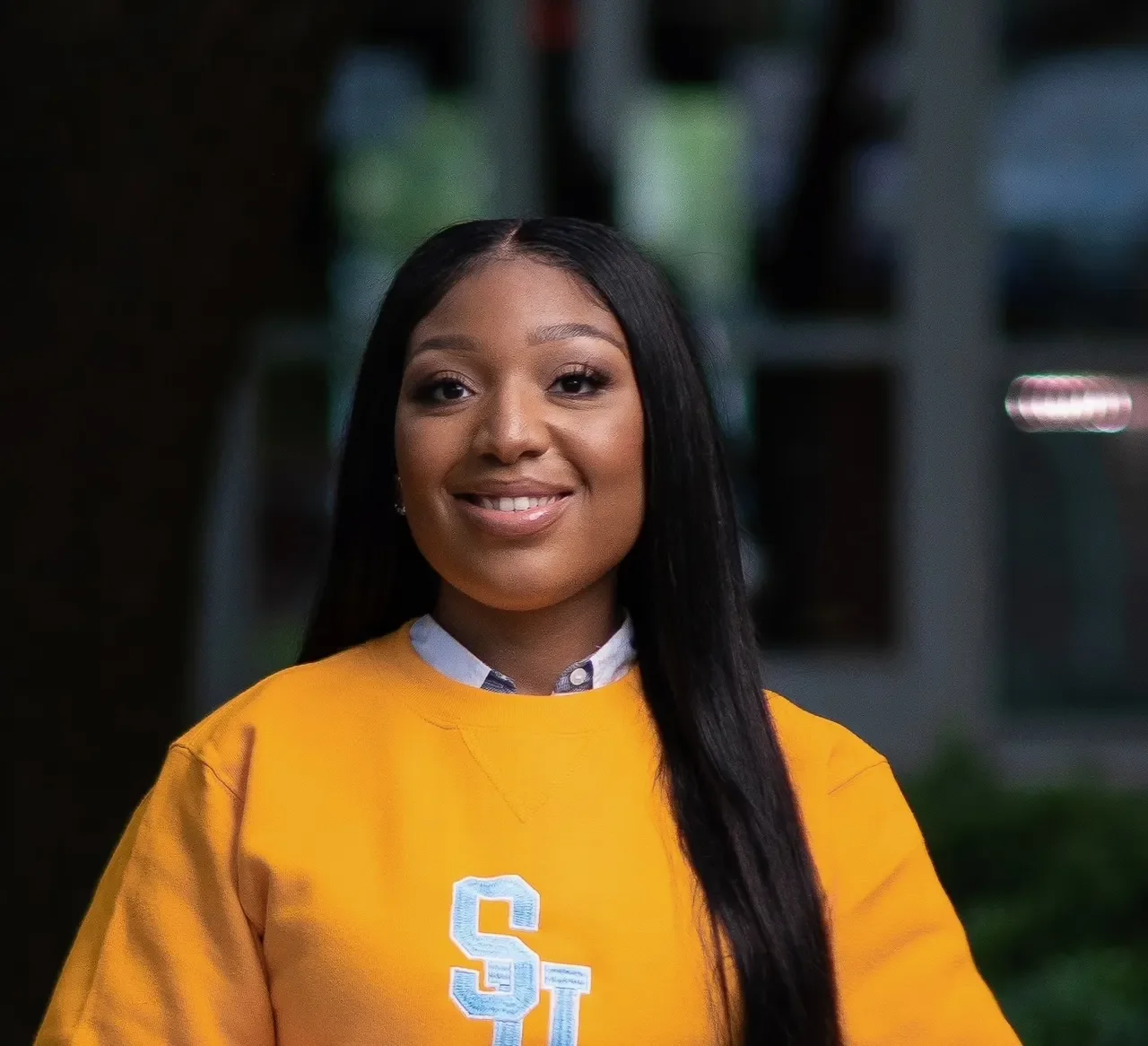 A young woman with long black hair smiling, wearing a yellow sports jersey with a white and blue collar and oversized letter 'S' on the front, standing outdoors with a blurred background of a building and greenery.