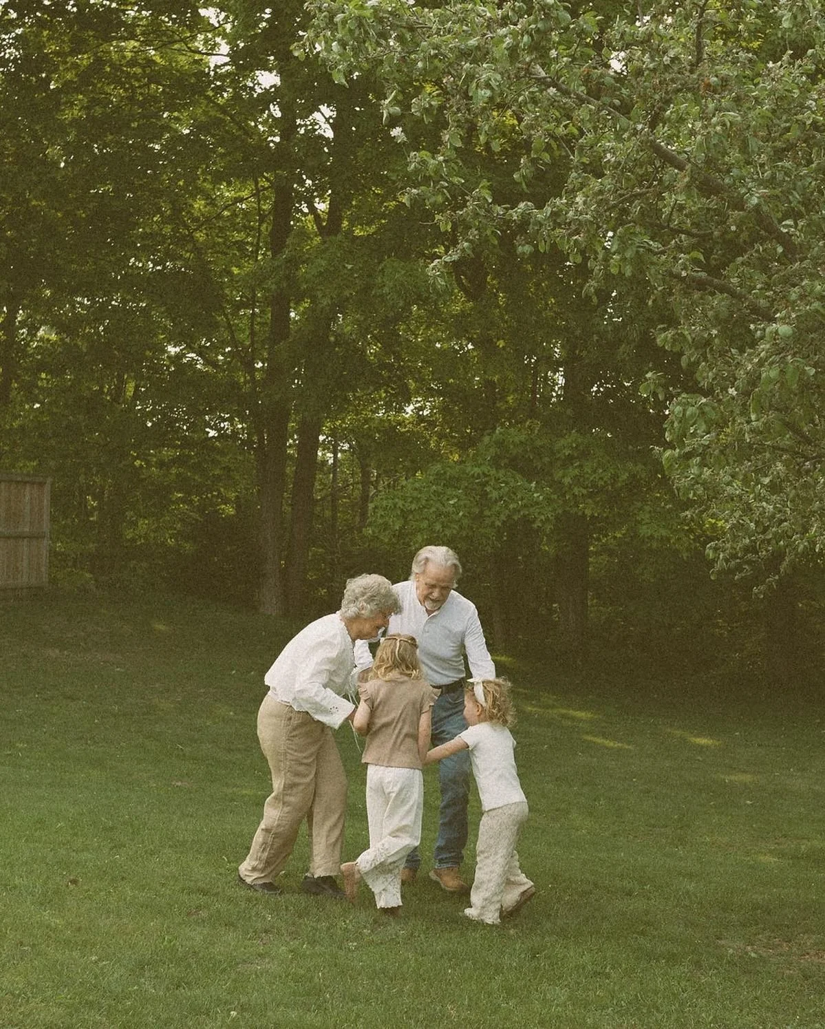Four elderly adults and two children playing together on a grassy lawn in a wooded backyard.