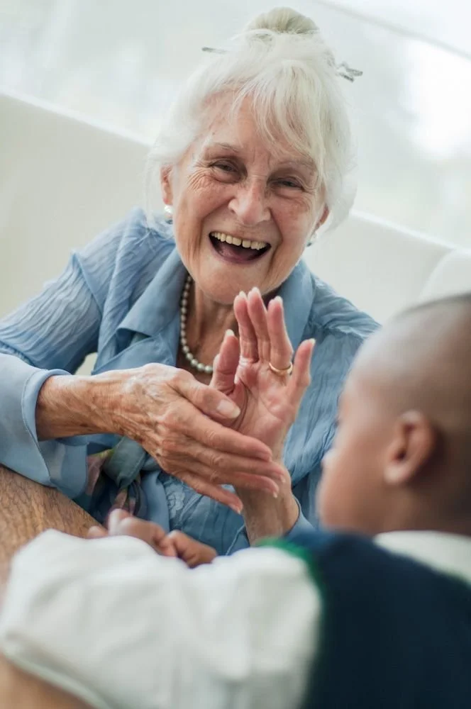 An elderly woman with white hair smiling and clapping hands with a young child in a hospital bed.