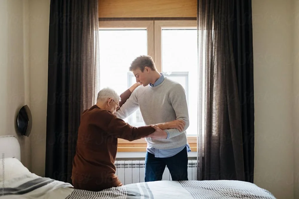 A young man helping an elderly woman stand up in a bedroom with sunlight streaming through a window behind them.