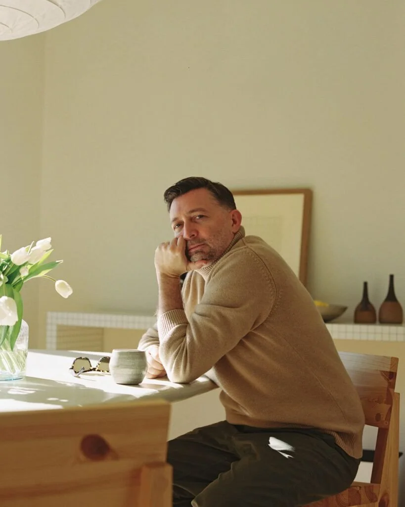 A man sitting at a dining table with his chin resting on his hand, looking towards the camera. The table has a vase with white tulips, a pair of sunglasses, and a small bowl. The setting appears to be a bright, modern kitchen or dining area with minimalist decor.