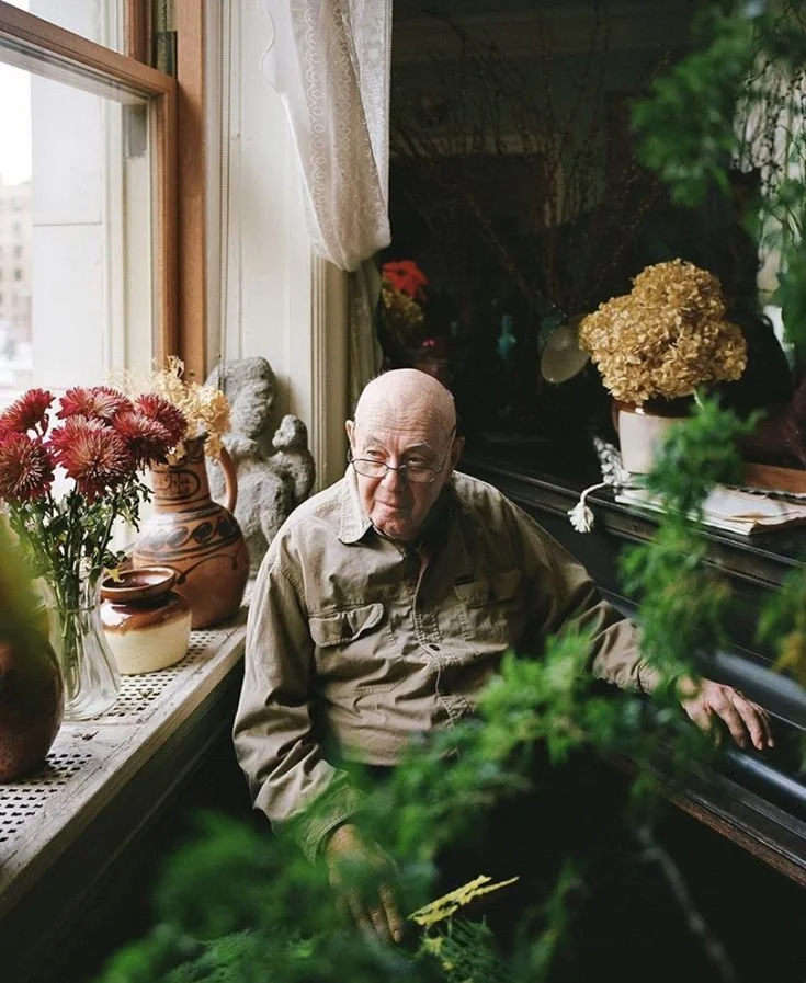 An elderly man with glasses sitting at a piano in a room decorated with several potted plants and vases of flowers, near a window with lace curtains.