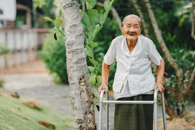 An elderly woman using a walker outdoors in a lush, green setting