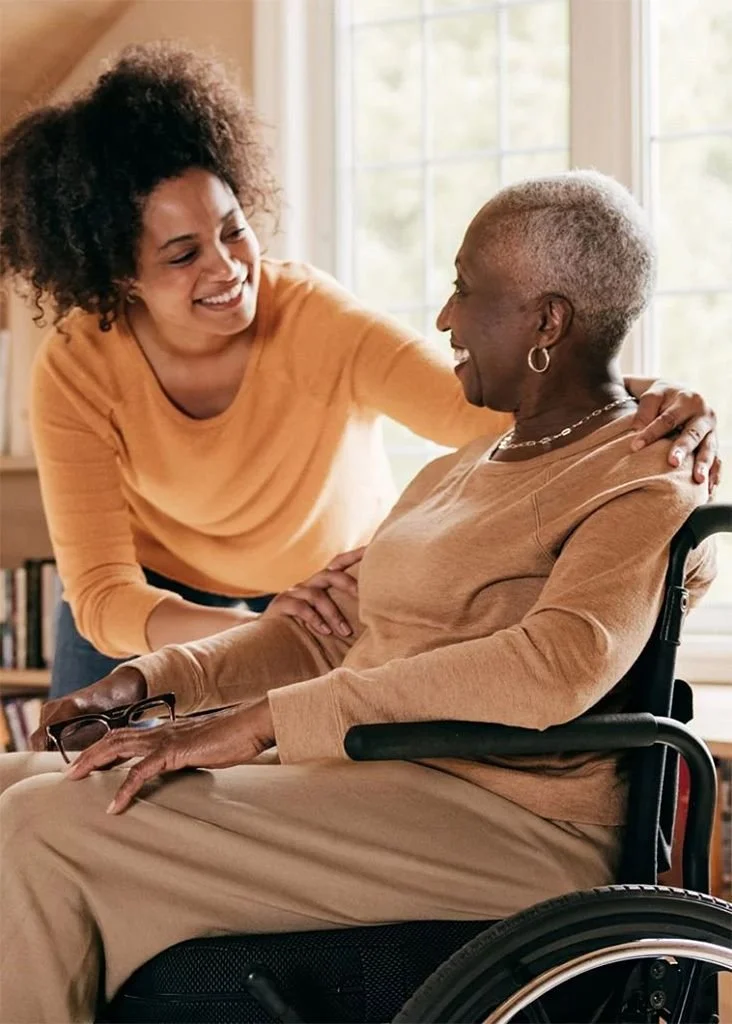 A woman with curly hair leaning towards an elderly woman in a wheelchair, smiling and engaging in conversation inside a well-lit room.
