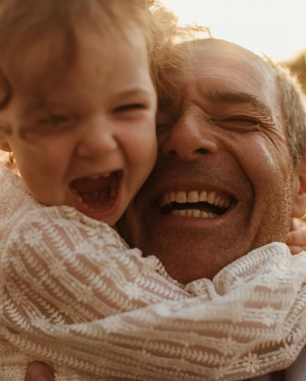 A joyful elderly man hugging a laughing young girl outdoors during sunset.