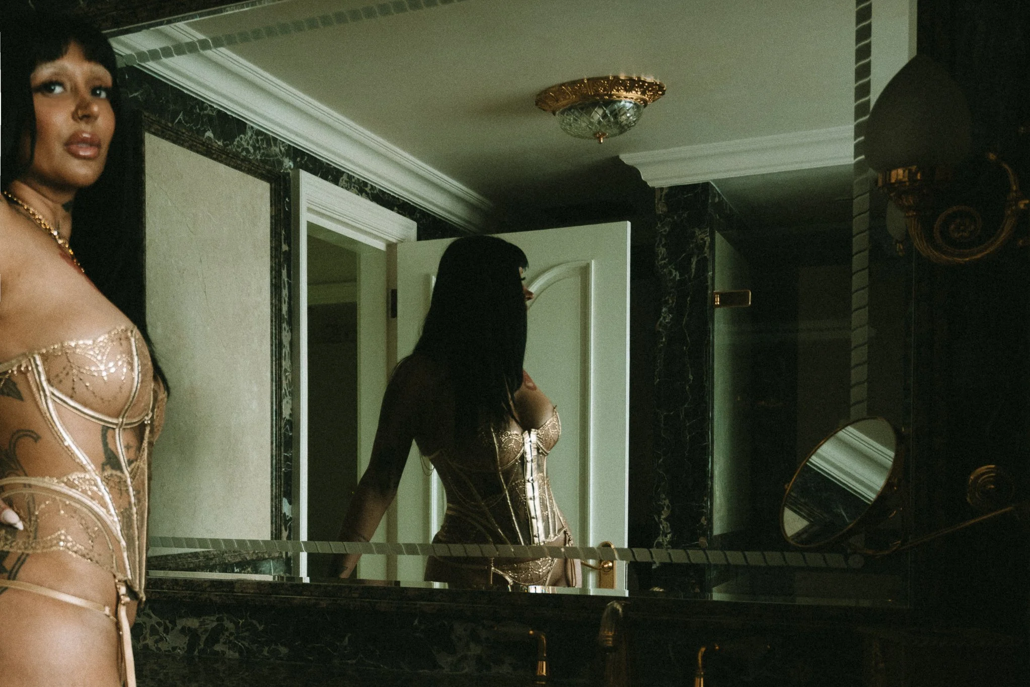 A woman with dark hair and tattoos wearing a beige corset with decorative details, standing in front of a mirror in a luxurious bathroom with black marble accents and gold fixtures.