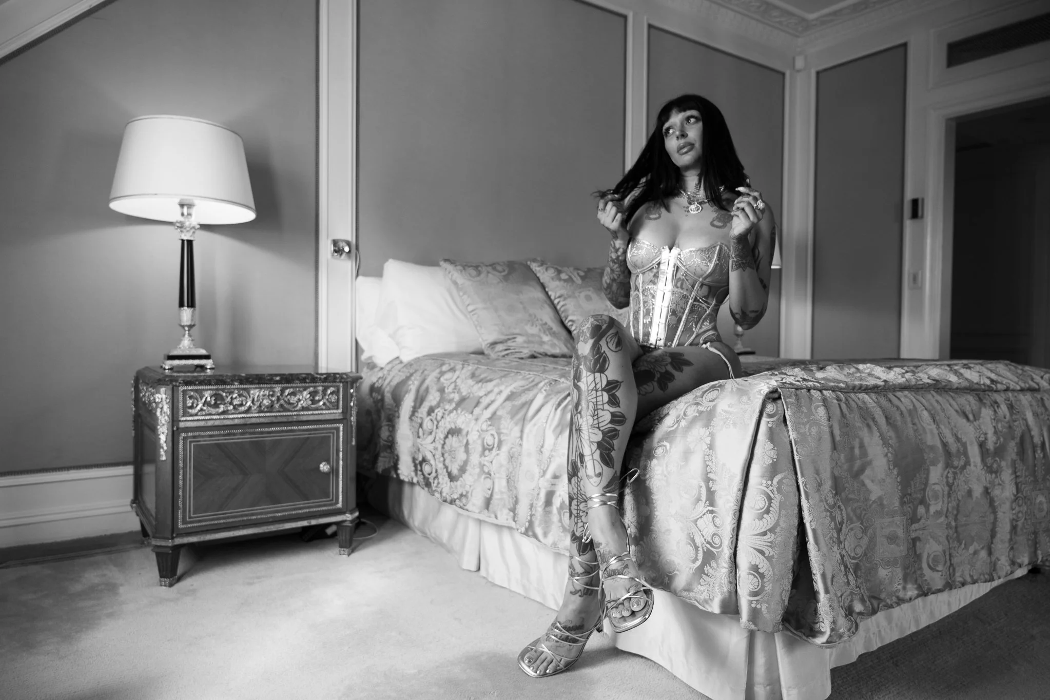 A woman with tattoos and long dark hair sitting on the edge of a bed in a hotel room, wearing a corset and high heels, holding a strand of her hair.
