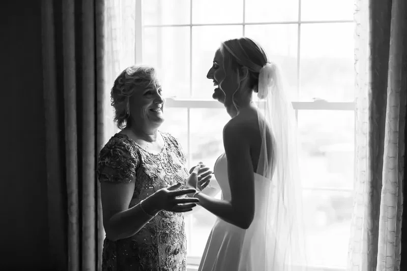 A bride and an older woman, possibly her mother, sharing a joyful moment by a window, with the bride in a wedding dress and veil, smiling and holding hands.
