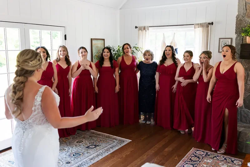 Bride in white dress with long blonde hair talking to nine bridesmaids dressed in matching red gowns in a cozy living room