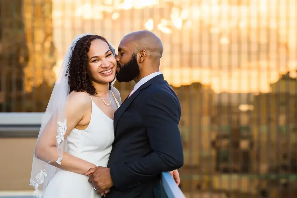 Bride smiles as the groom kisses her cheek during a romantic rooftop wedding portrait at sunset.