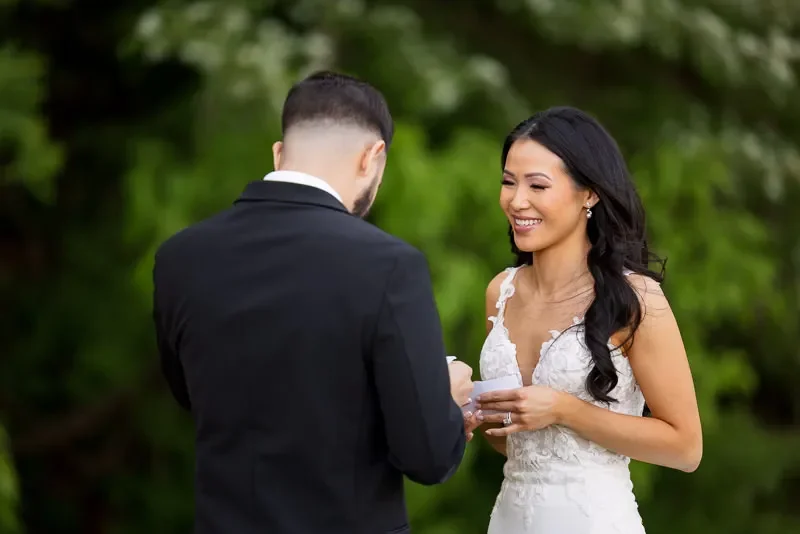 A bride in a white wedding dress and a groom in a black suit exchanging rings outdoors.