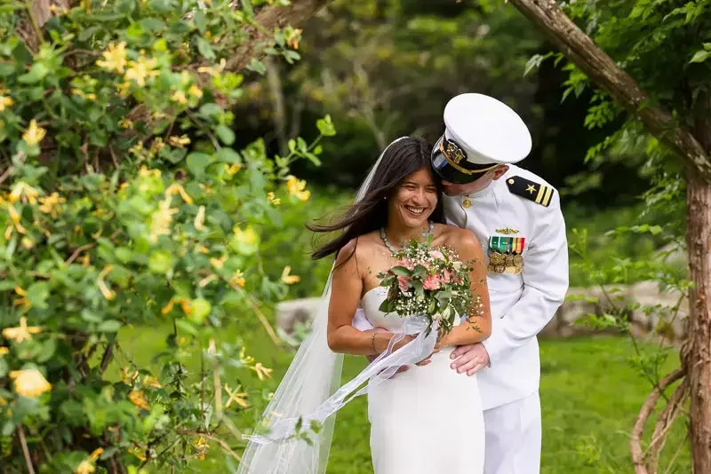 Bride smiling as groom in a naval uniform embraces her from behind during an outdoor wedding portrait surrounded by greenery.