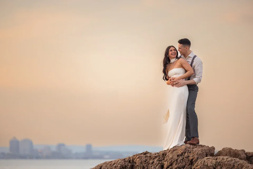 Engaged couple embracing on rocky shoreline during sunset