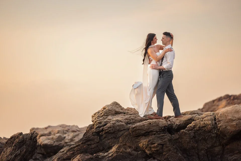 Engaged couple embracing on rocks with flowing dress at sunset