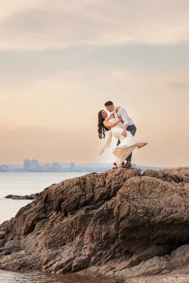 Engaged couple embracing on rocky shoreline at sunset during coastal engagement session.