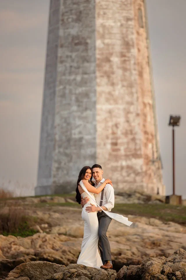 Engaged couple hugging in front of a lighthouse during seaside engagement session.