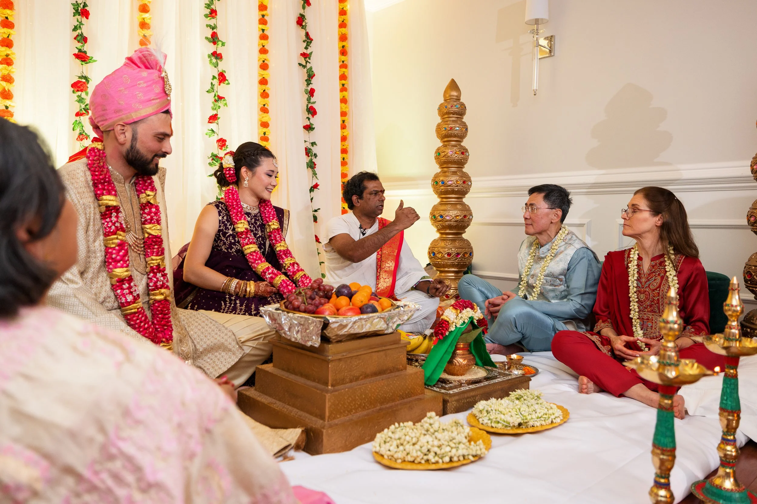 A couple adorned with garlands sits facing a priest during a ceremony.
