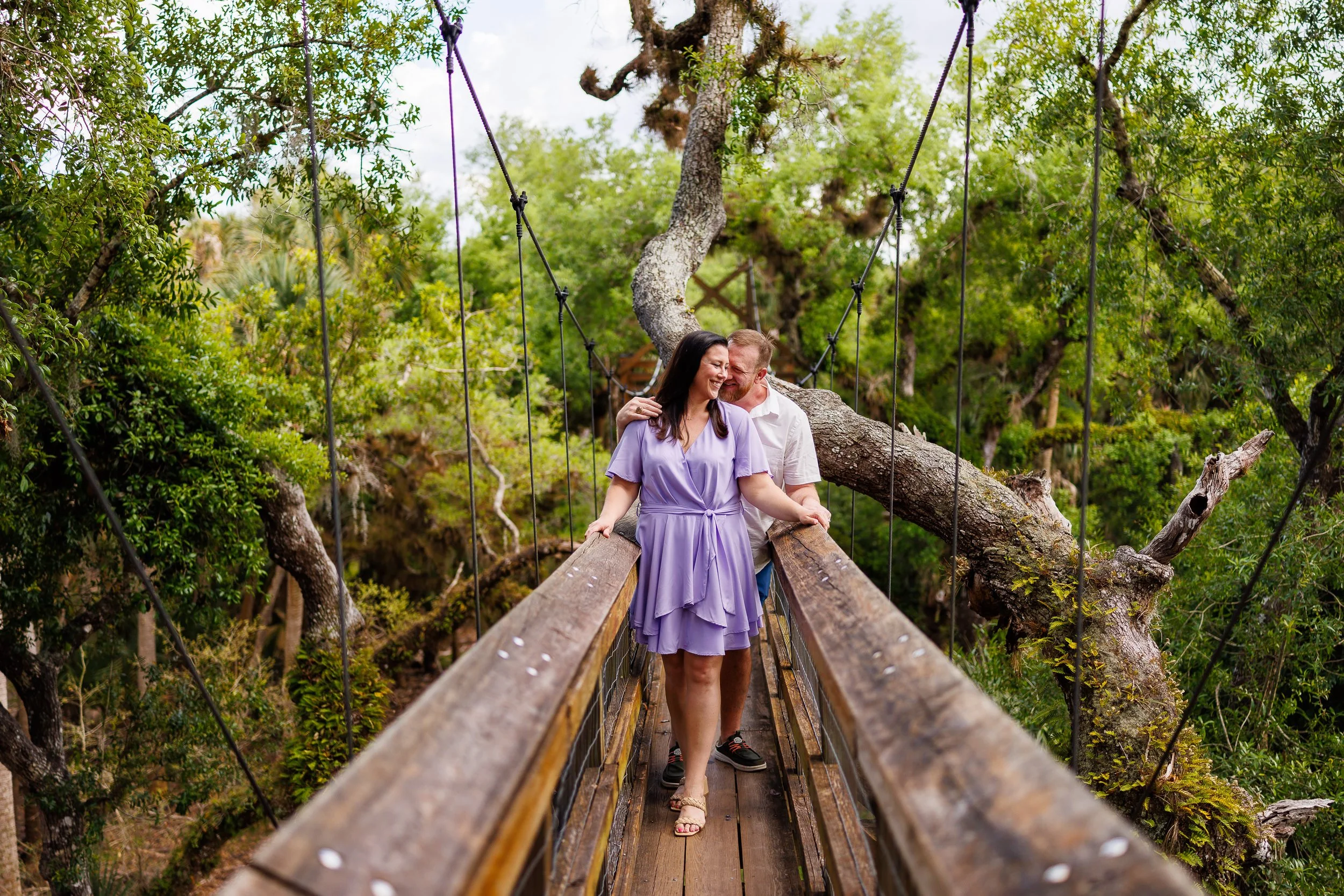 Will and Erin partway across the Myakka canopy walkway, the bridge rails running away toward the oak canopy behind them.
