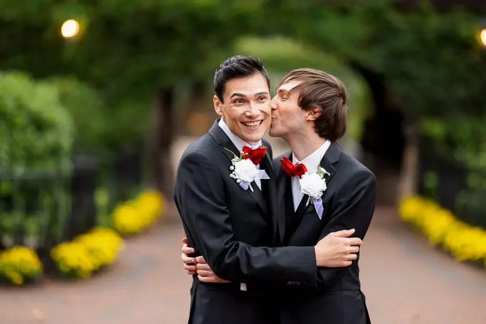 Two men in wedding tuxedos embracing and kissing in a park, with greenery and yellow flowers along a pathway in the background.
