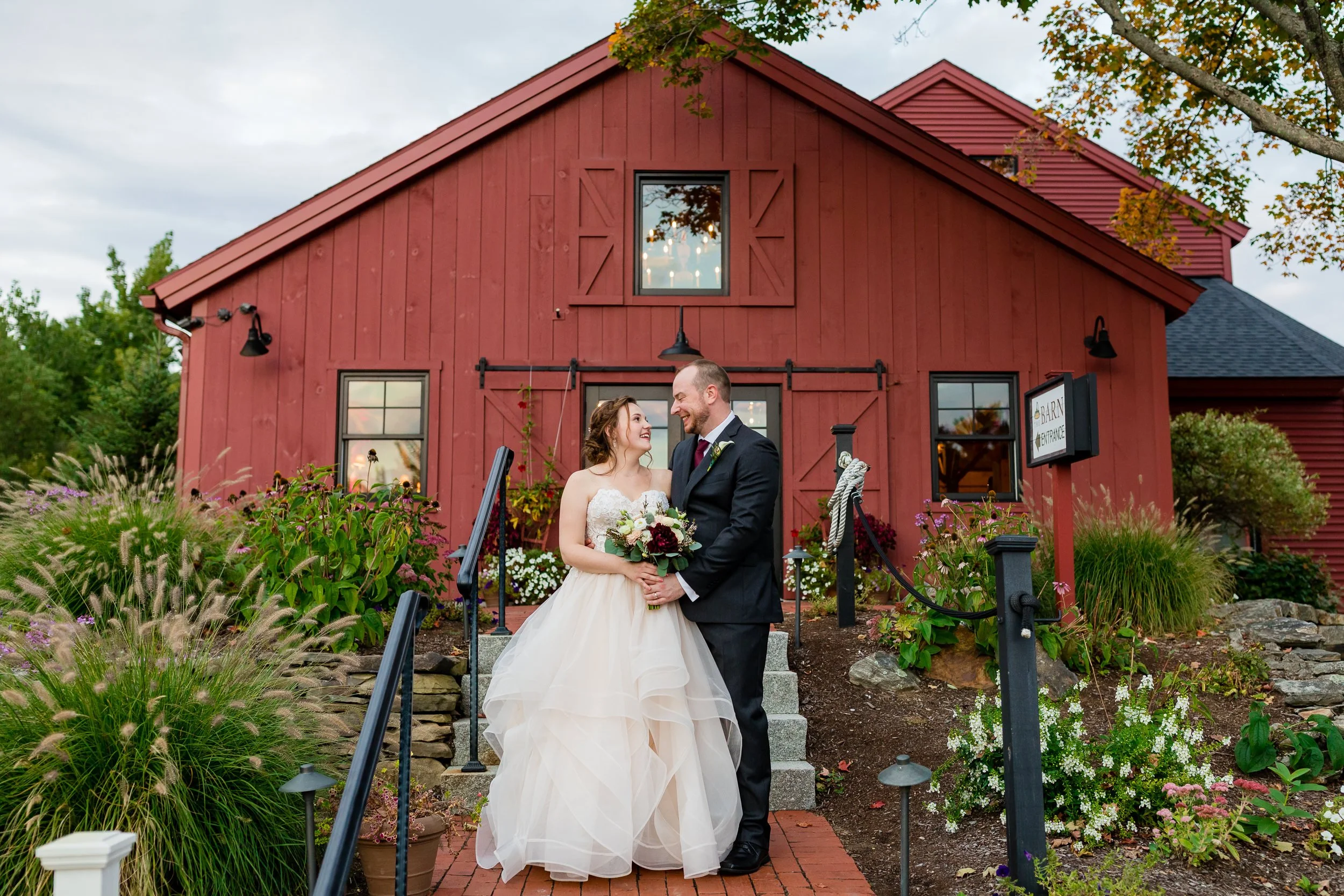 A bride and groom outside a red New England barn, autumn foliage and ornamental grasses around them.