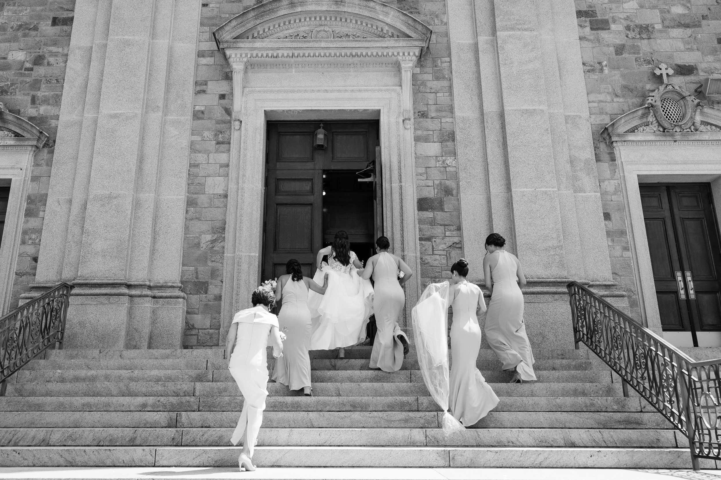 Bridesmaids in white gathering their dresses and hurrying up the stone steps of a church behind the bride.
