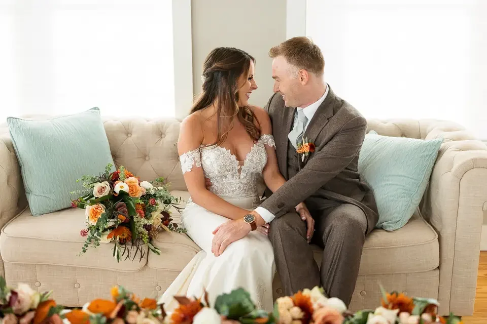 A bride and groom sit close together on a beige sofa, smiling and looking into each other's eyes. The bride wears a white lace wedding dress, and the groom wears a gray suit with a boutonnière. A bouquet with orange, peach, and white flowers rests on the sofa next to them.