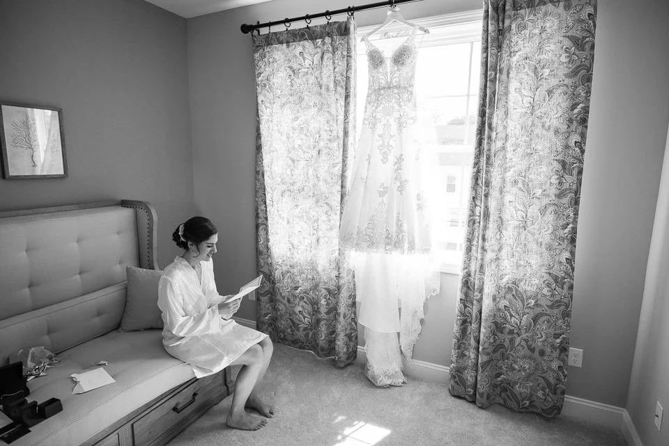 A person in a white robe sits on a bed, looking at a dress hanging by a window.