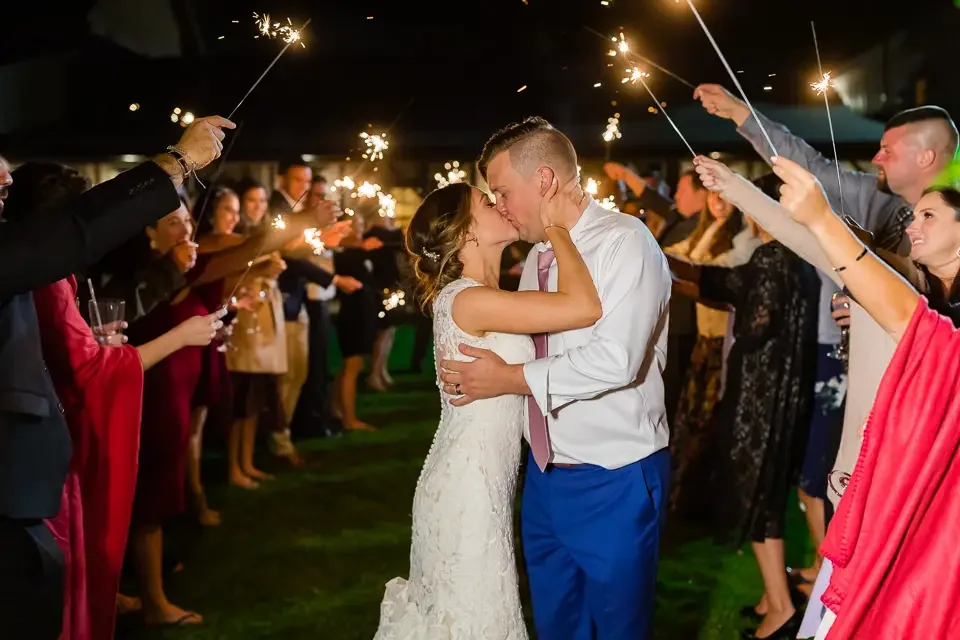 Newlyweds share a kiss during a joyful sparkler exit surrounded by wedding guests at night.