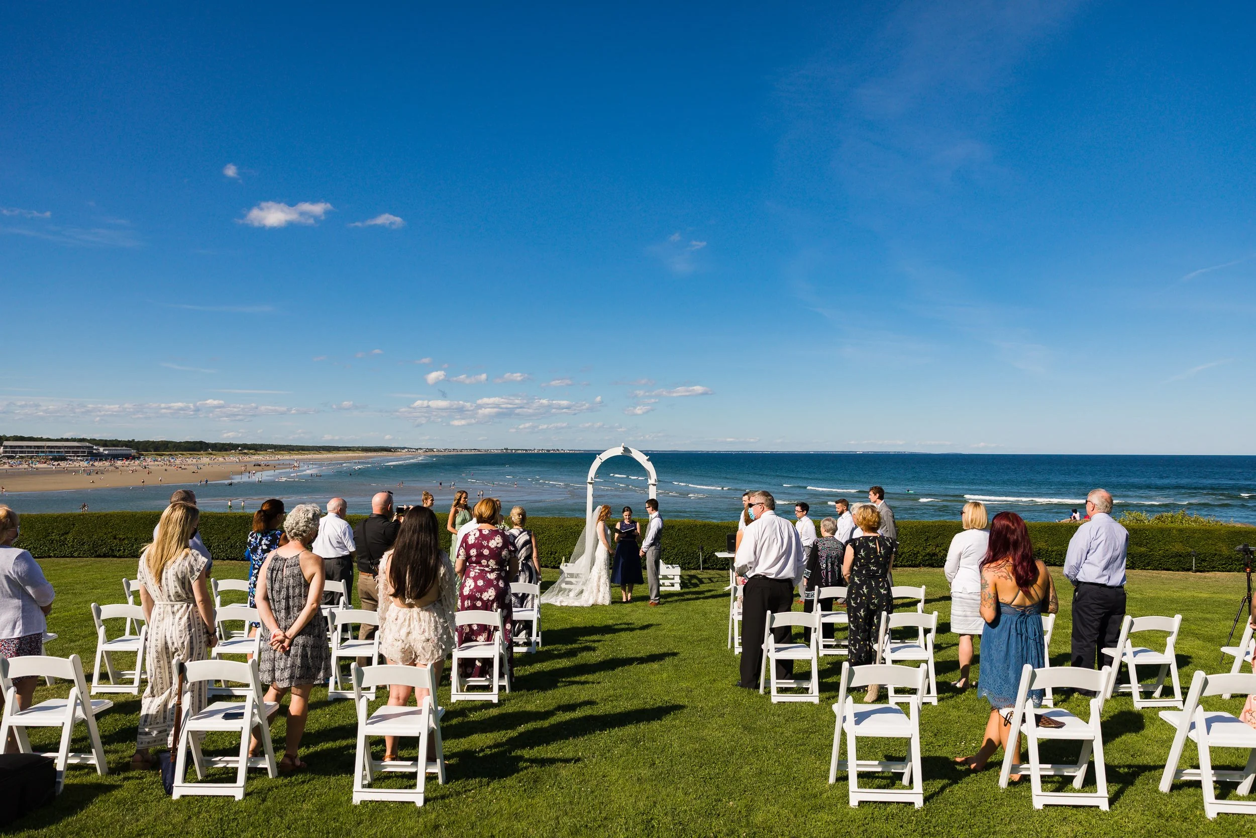 A wedding ceremony on a lawn above the ocean in Maine, guests standing as the couple exchange vows.