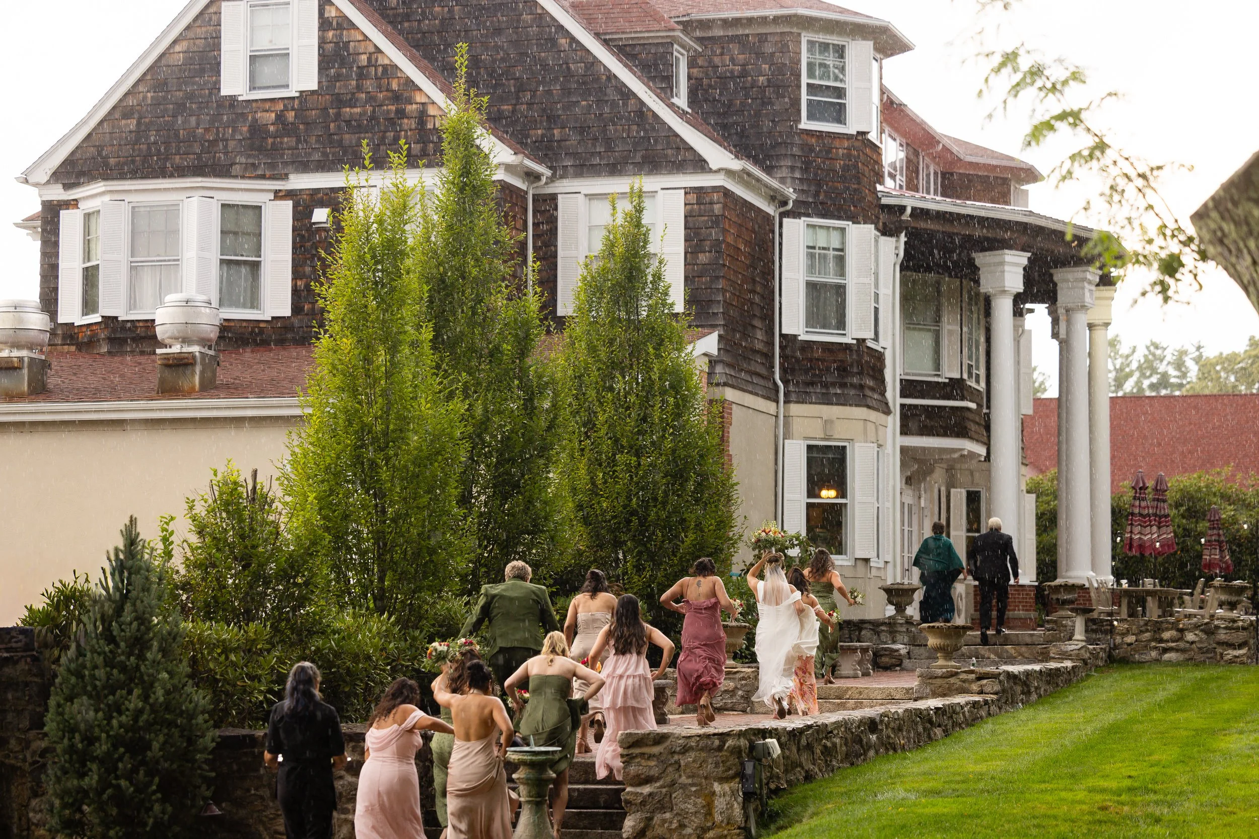 Wedding guests and the bridal party hurrying up stone steps toward a large shingled house in visible rain.