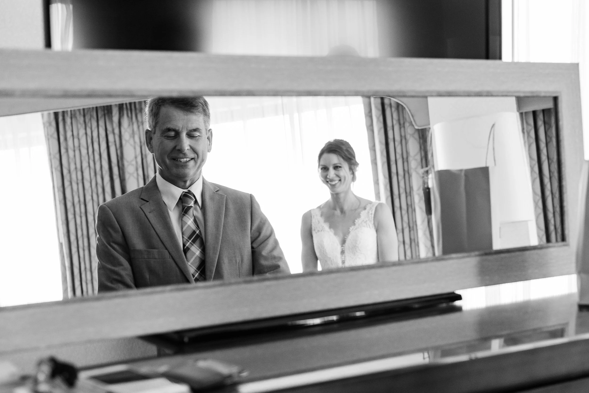 A father seen in a mirror, looking down, with his daughter in her wedding dress reflected behind him.