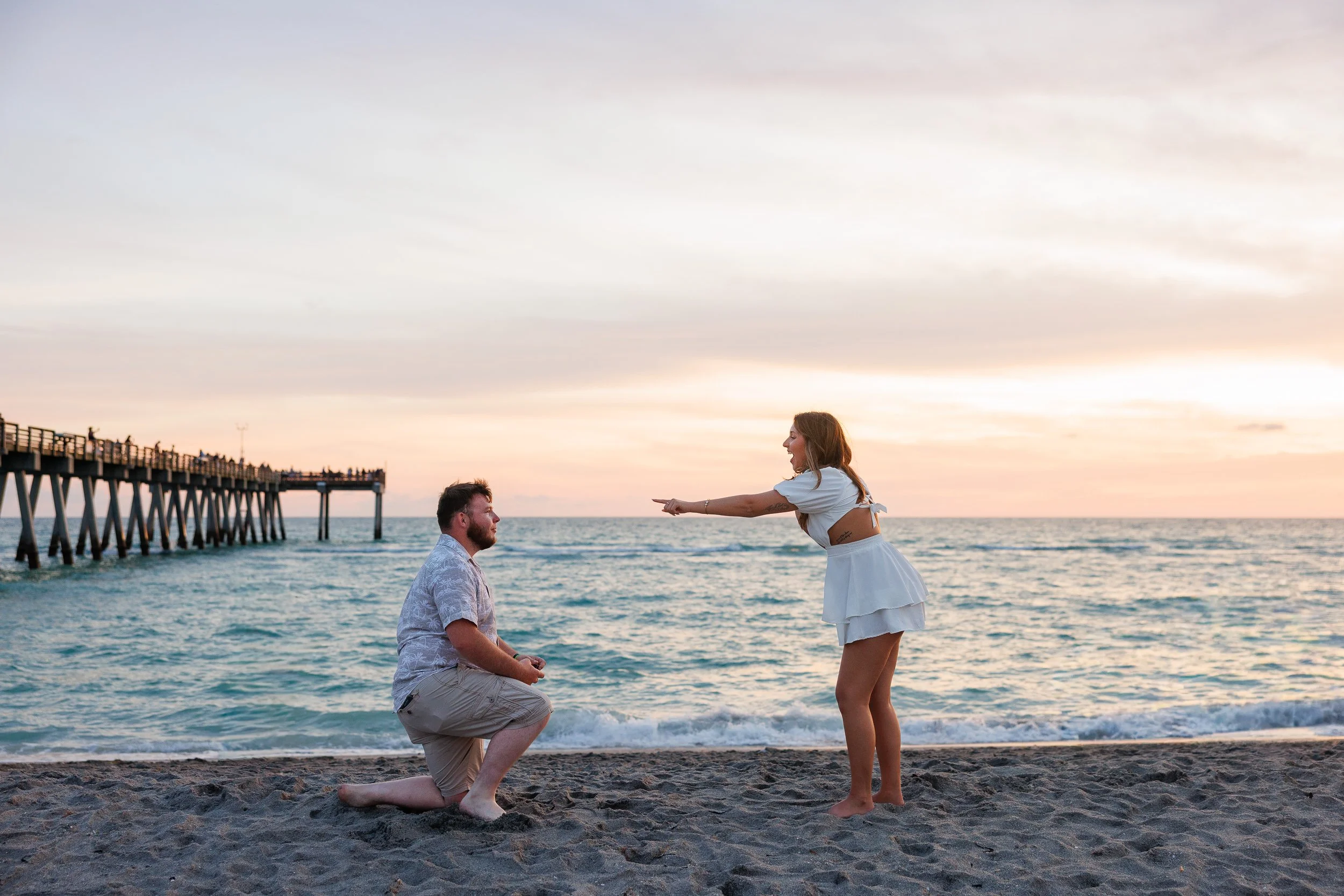 A man on one knee proposing on the sand at Venice Beach, the fishing pier behind him and the Gulf turning pink at sunset.