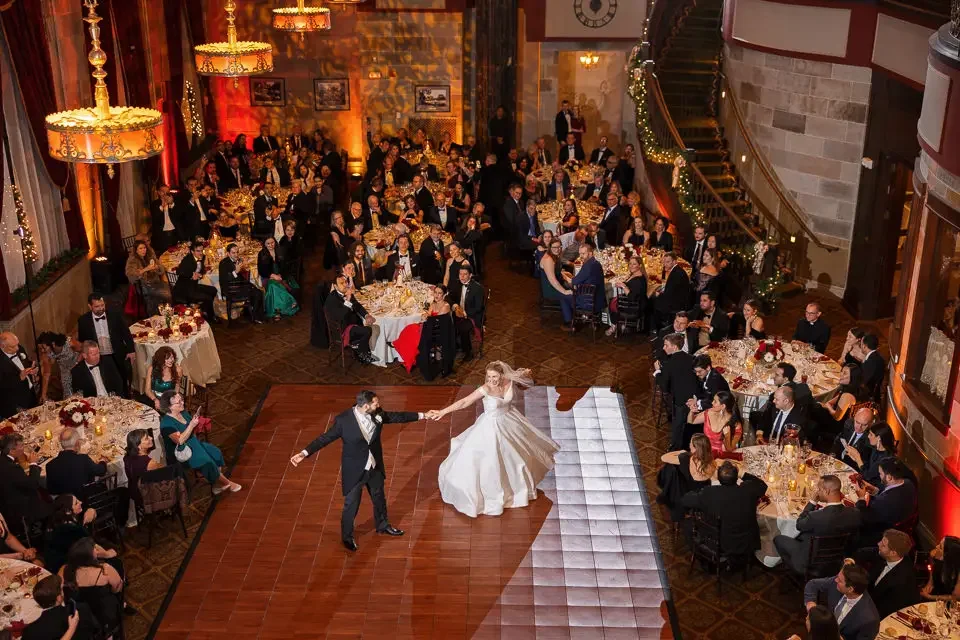 A bride and groom are dancing together on a dance floor at a wedding reception, surrounded by seated guests at decorated tables in a large, ornate hall.