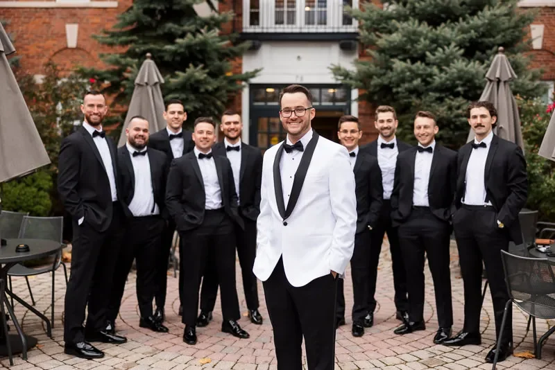 Group of eight men in tuxedos, with one man in a white tuxedo jacket standing in front, outdoors with trees and umbrellas.