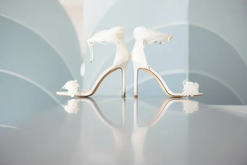 White high-heeled shoes with lace and feather details, positioned to look like a pair of symmetrical wings, reflected on a shiny surface.