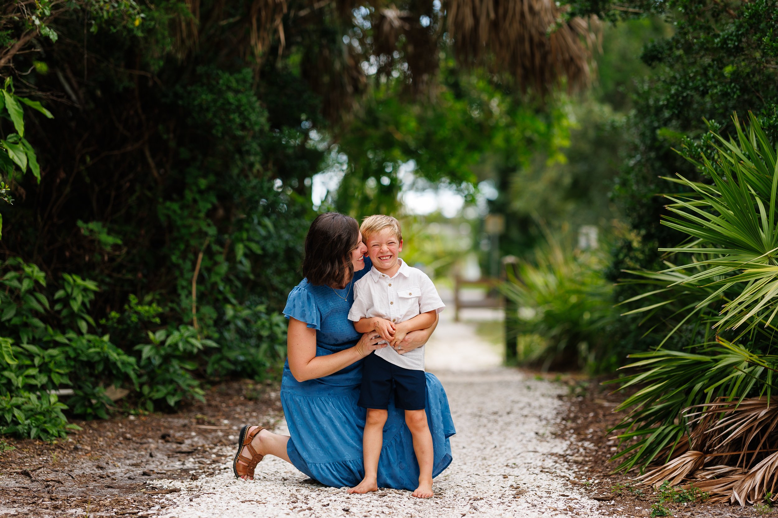 A person in a blue dress holds a child on a gravel path surrounded by greenery.