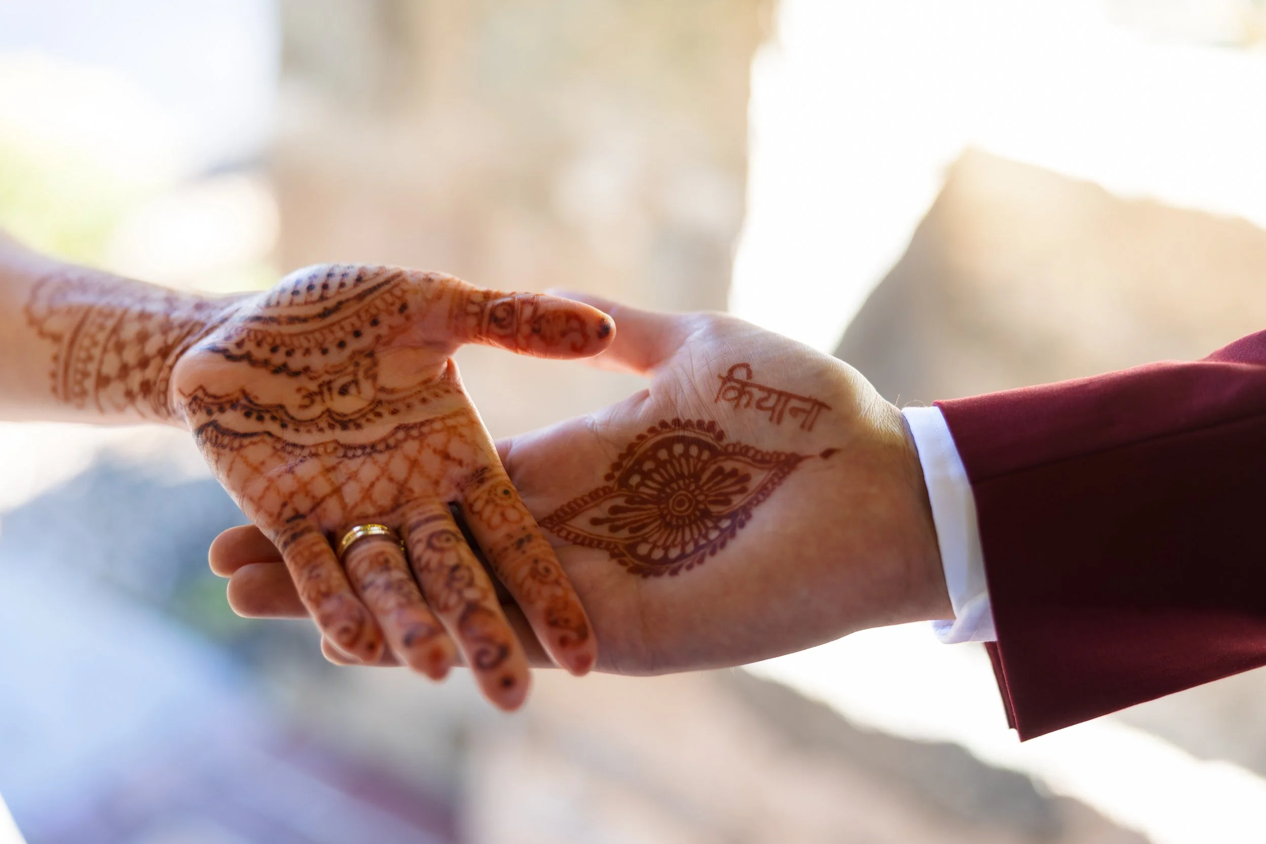 Two hands with henna and rings forming a heart shape, backlit against a bright background.