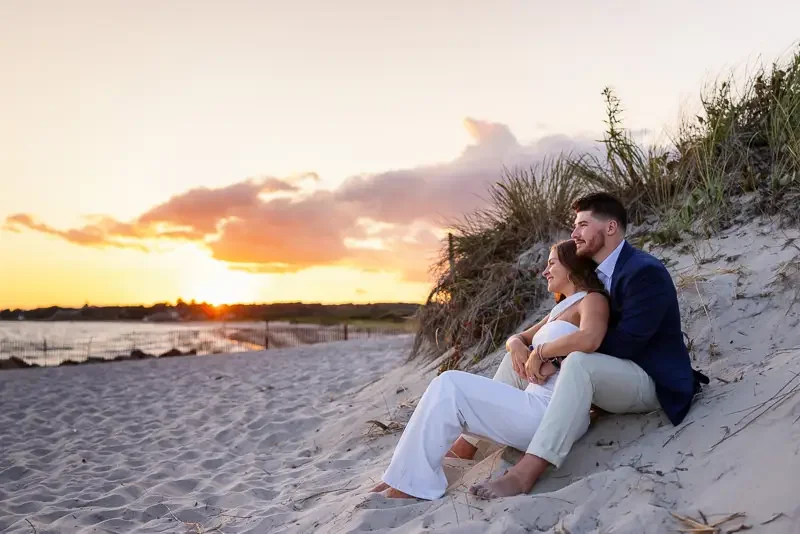 A couple sitting on sand dunes at sunset, embracing with a scenic beach and water in the background.