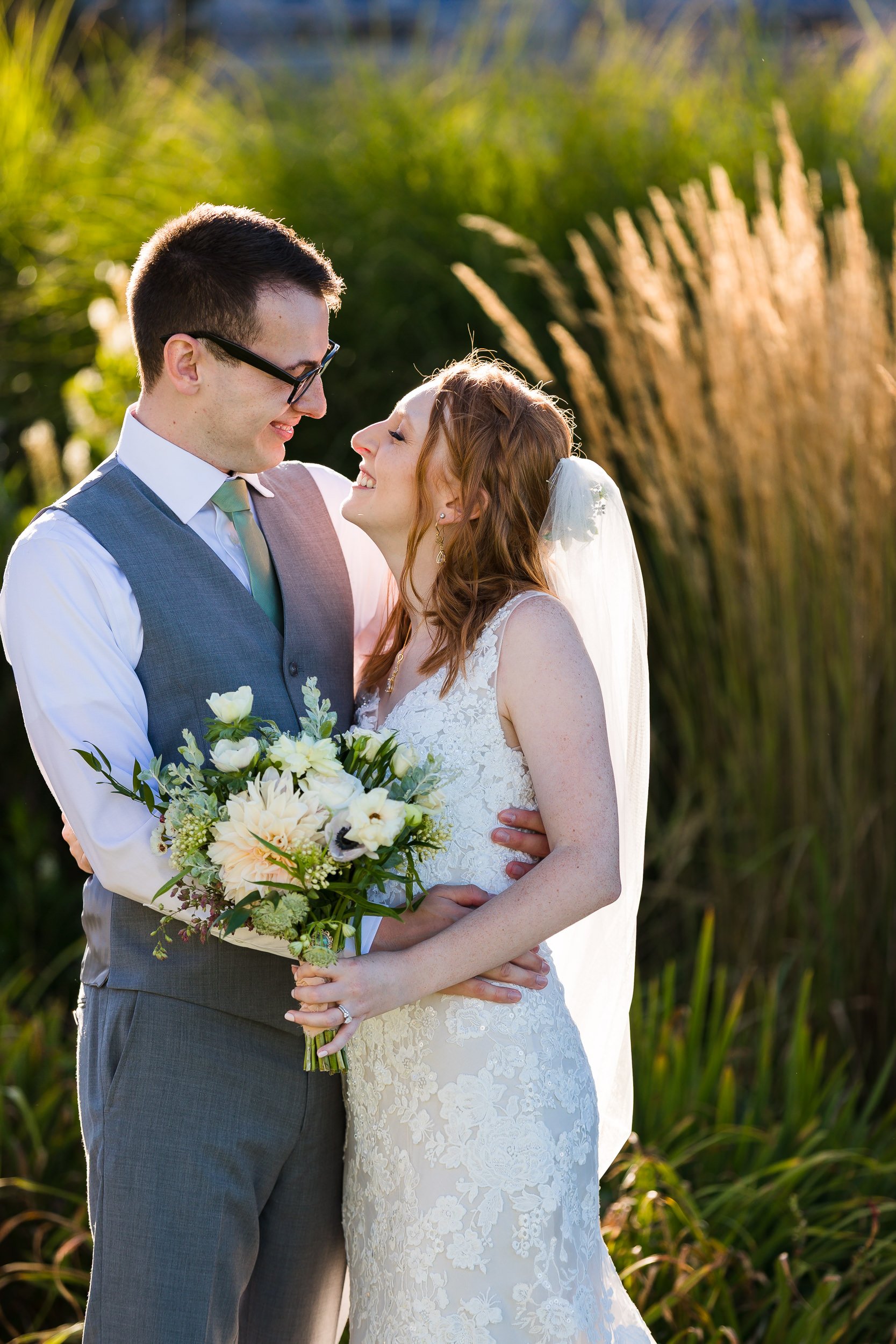 A bride laughing up at her groom in tall ornamental grasses, late afternoon light behind them.