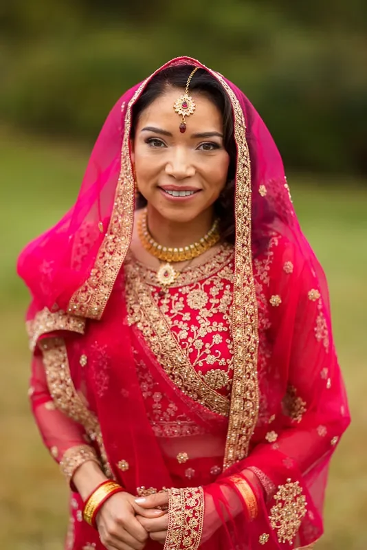 A woman in traditional Indian bridal attire, wearing a red saree with gold embroidery, jewelry, and a pink veil, standing outdoors.