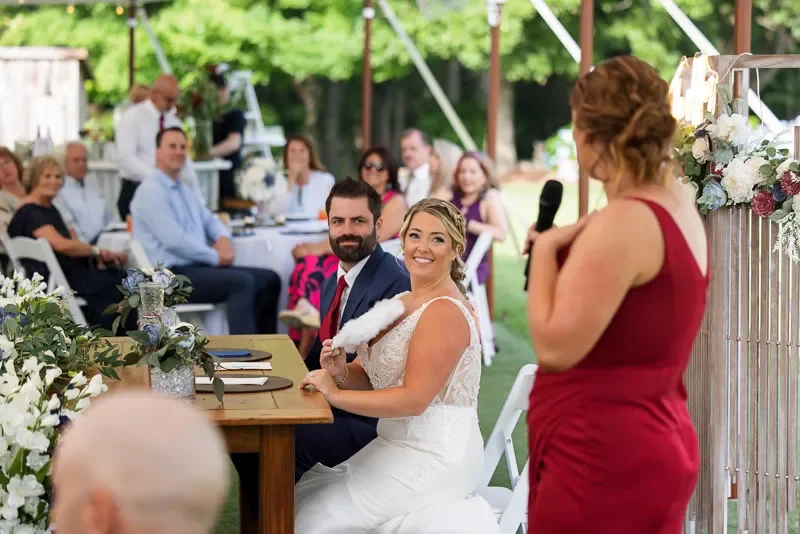 Bride and groom sitting at a table during their outdoor wedding reception, listening to a woman in a red dress giving a speech with a microphone.