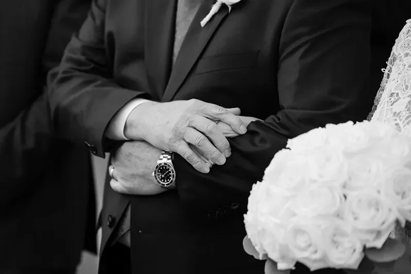 Black and white photo of a person in a suit crossing their arms, with a wristwatch visible, standing next to a bride holding a bouquet of roses.