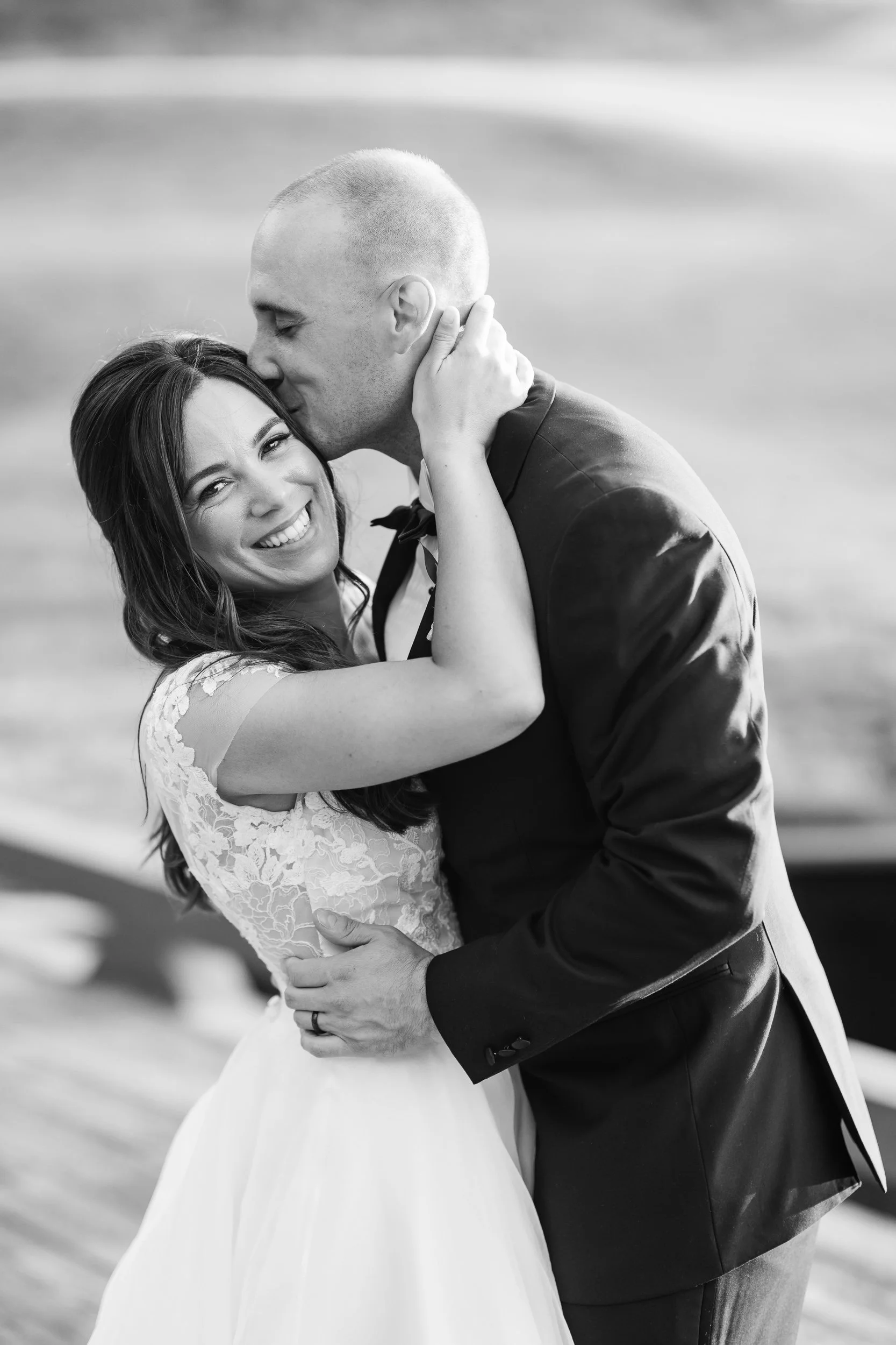 A groom kissing his bride's temple while she smiles at the camera, close, in black and white.
