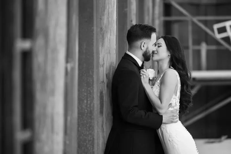 A black and white photo of a bride and groom close together, leaning against a wooden wall, about to kiss, with the groom in a tuxedo and the bride in a lace wedding dress.